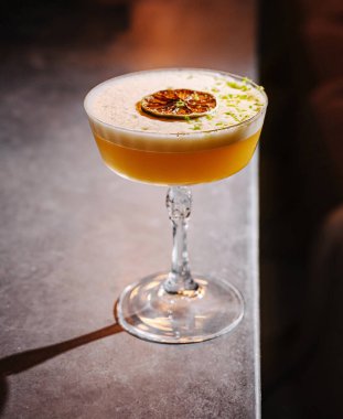 Güzelce hazırlanmış bir kokteyl bir bar tezgahının üzerinde köpüklü kahverengimsi sarı bir renk ve kurutulmuş bir narenciye ile duruyor. Ortam, içkinin sofistikesini vurguluyor..