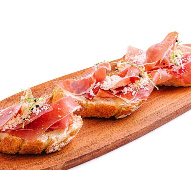 Üç parça bruschetta, üzerinde prosciutto ve peynir serpiştirilmiş ahşap bir tepside oturur. Yemek zarifçe sunuluyor, tazelik ve lezzeti vurguluyor..