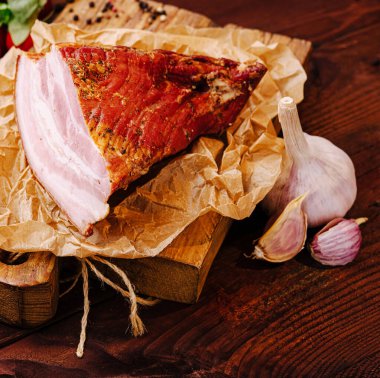 Füme jambonun bir kısmı, buruşuk parşömenle çevrili ahşap bir kesme tahtasının üzerinde dinleniyor. Taze sarımsak karanfilleri sunuma kırsal bir dokunuş ekler..
