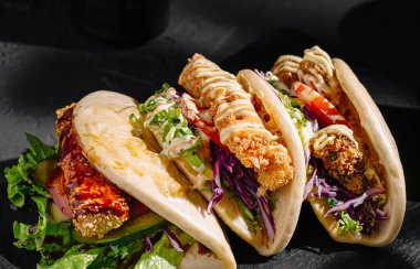 Üç kızarmış tavuk sandviçi, taze marul, doğranmış lahana ve üzerine sos serpiştirilmiş siyah bir tabağa konur. Yemekler çeşitli malzemeler ve tatlar sergiliyor..