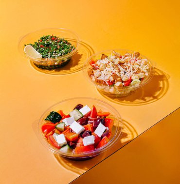 Canlı turuncu bir yüzey üzerinde üç farklı salata düzenlenir. Her salatada taze sebzeler, bitkiler ve peynir var. Renkli bir karışım sergiliyorlar. Sağlıklı bir yemek için mükemmel..