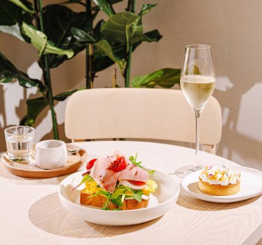 Bir brunch masasında yeşil ve jambonlu renkli bir sandviç, yanında tatlı bir pasta ve bir bardak köpüklü şarap bulunur..