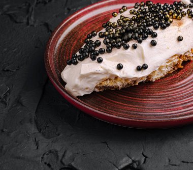 Bir gurme tatlıda siyah incilerle süslenmiş yuvarlak kırmızı bir tabağa zarif yerleştirilmiş kremalı bir tepsi bulunur. Yemek pişirme sanatını ve sofistikeliği yansıtıyor..