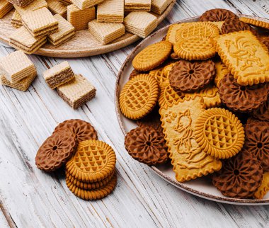 Tabaklara yerleştirilmiş farklı tipte kurabiyeler ahşap bir masada duruyor. Çeşitli gofretler, kremalı kurabiyeler ve yuvarlak bisküviler içeriyor..