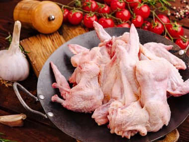 Bir tepsi çiğ tavuk kanadı ahşap bir masada taze domates ve sarımsakla çevrilidir. Malzemeler lezzetli bir pişirme işlemi için hazırlandı..