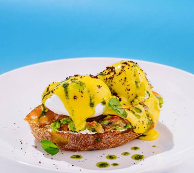 İki mükemmel haşlanmış yumurta, avokado ve somonla kızartılmış ekmeğin üzerinde, kremsi sarı bir sosla serpilmiş ve parlak mavi bir zemin üzerinde taze otlarla çevrili..