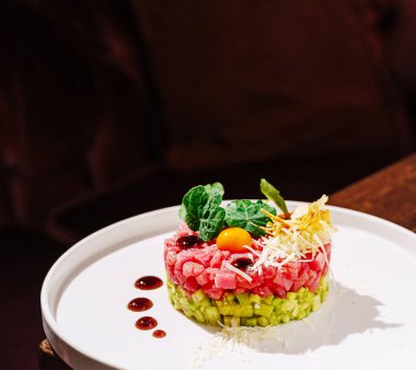 Renkli bir suşi tartar sofistike bir yemek ortamında beyaz bir tabakta sunulur. Ton balığı, avokado ve garnitürleri rendelenmiş..