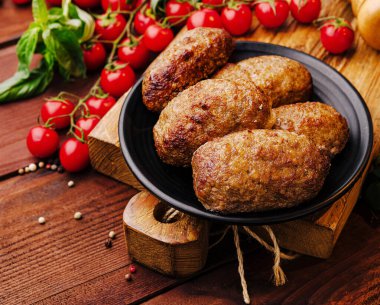 Ev yapımı köfteler siyah bir tabakta otururlar, kırsal bir ahşap yüzeyde parlak kırmızı kiraz domatesleriyle çevrili. Yemek taze malzemeleri ve geleneksel yemekleri ön plana çıkarıyor..