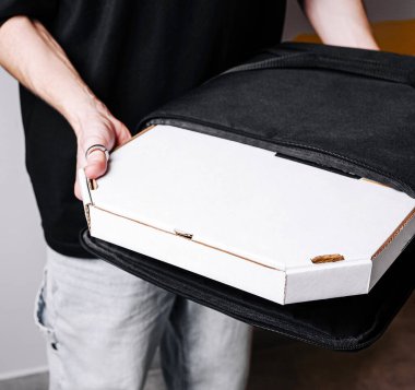 Modern bir kapalı mekan ortamında bir kişi pizza kutusunu siyah bir teslimat çantasının içinde dikkatlice tutar. Teslimata hazır olan kutuya odaklanıyoruz. Tipik bir yemek servisi anı sergiliyoruz..