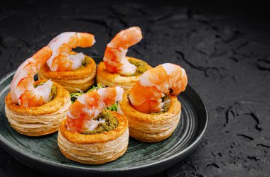 A plate features five golden pastry shells topped with cooked shrimp and green filling. The savory appetizers are displayed against a dark background, highlighting their colors. clipart