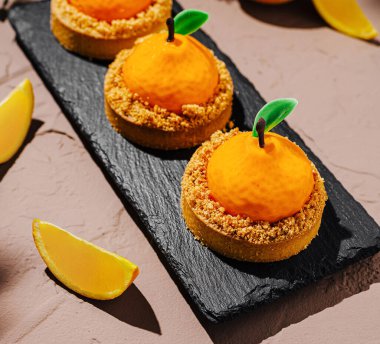 Sanatsal olarak dizilmiş turunçgillerden esinlenilmiş tatlılar parçalanmış bir tabanın üzerinde canlı turuncu küreler içerir. Çevreleyen limon dilimleri zarif sunuma ferahlatıcı bir dokunuş ekler.