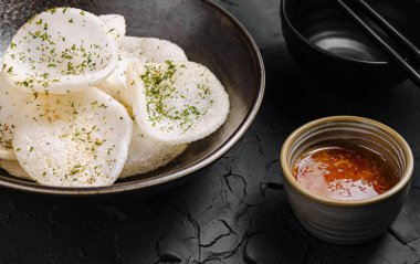 A bowl filled with crispy rice crackers, garnished with herbs, accompanies a small cup of spicy dipping sauce. The textured dark background enhances the vibrant colors. clipart