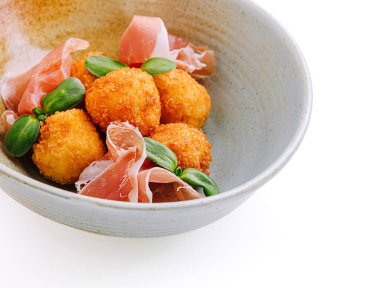 Kızarmış kroketler bir kasede sunulur, jambon dilimleri ve taze yeşilliklerle süslenir. Yemekler leziz bir lezzet ve doku kombinasyonu sergiliyor..