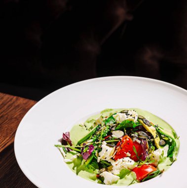 Yeşillikler, kiraz domatesleri, tohumlar ve otlar içeren canlı bir salata modern bir restoran ortamında zarif bir şekilde sunulur, sağlıklı yemek seçenekleri sunar..