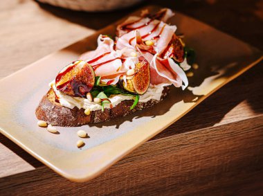 Güzel düzenlenmiş açık yüzlü bir sandviçte taze incir, jambon dilimleri ve çam fıstığıyla süslenmiş krem peynir yer alıyor. Sıcak aydınlatma yemeğin cazibesini arttırır..