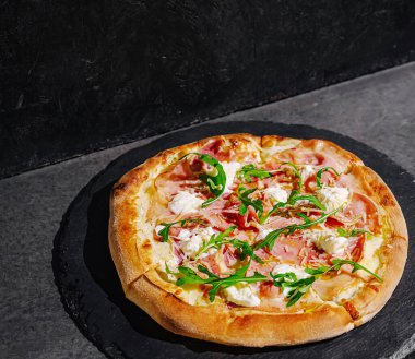 Lezzetli bir pizza siyah taştan bir yüzeye konur, jambon, krem peynir ve canlı roka içerir. Kabuğu altın ve çıtır çıtır, cezbedici bir zevk..