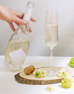 Bir el desenli bir şişeden köpüklü bir içeceği uzun bir bardağa doldurur. Taze çiçekler ve bir mantar kırsal ahşap yüzeyi süslüyor, kutlama için zarif bir ortam yaratıyor..