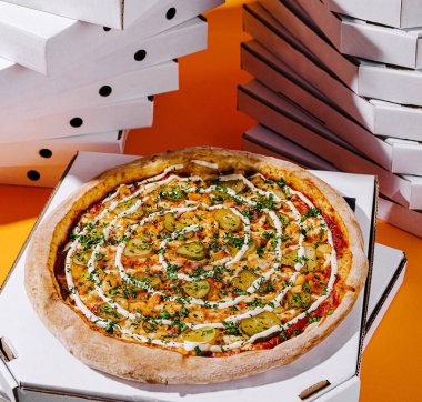 A fresh pizza topped with ingredients like pickles, chicken, and herbs is showcased in front of a vibrant orange background and a stack of white pizza boxes. clipart