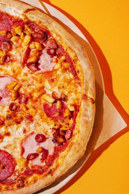 Yuvarlak pizza pepperoni, jambon ve ananas ile kaplıdır, ahşap bir tahta üzerinde, parlak sarı bir zemin üzerinde dinlenir, lezzetli malzemelerini vurgular ve davetkar bir görünüm sunar..