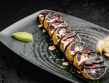 Taze suşi ruloları koyu dokulu bir tabakta ustalıkla düzenlenir. Wasabi, zencefil ve sosla süslenmişler, canlı bir sunum sergiliyorlar..