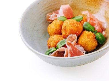 Bir kasede ince dilim prosciutto ve taze yeşil yapraklarla birlikte kızarmış altın toplar yer alıyor. Yemek zarif bir şekilde sunuluyor, kumaş ve renkler sergileniyor..