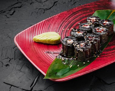 Lezzetli suşi ruloları çarpıcı bir kırmızı tabakta güzelce düzenlenir. Her rulo susam tohumuyla kaplanmış, yanında bir tutam wasabi, görsel olarak çekici bir sunum yaratıyor..