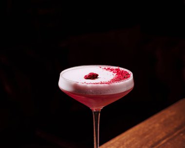 Canlı bir ahududu kokteyli, üzerinde ahududu tozu ve tek bir dut bulunan parlak bir bardakta sergilenir. Yumuşak ortam ışığı, gölgeli bir arkaplanda içkinin zengin rengini arttırır..