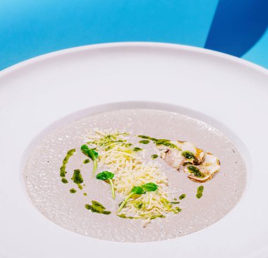 Peynir ve taze otlarla kaplı beyaz bir kasede yumuşak ve kremalı bir mantar çorbası servis edilir. Canlı bir arka plan modern yemek deneyimini geliştirir..