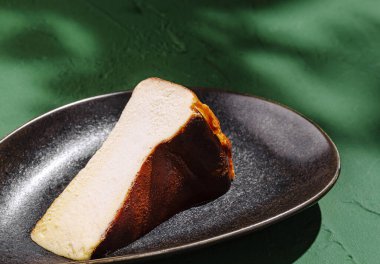 Yumuşak, kremsi bir dokusu ve koyu bir dış katmanı olan bir dilim peynirli kek, sığ, oval bir tabakta güzelce duruyor. Yeşil arkaplan sunuma canlılık katarak çekiciliğini artırıyor.