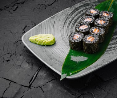 Renkli suşi ruloları, bir tutam wasabi ve dekoratif yaprak eşliğinde dokulu bir tabakta güzelce düzenlenir. Ayar, çağdaş bir yemek atmosferini yansıtıyor..