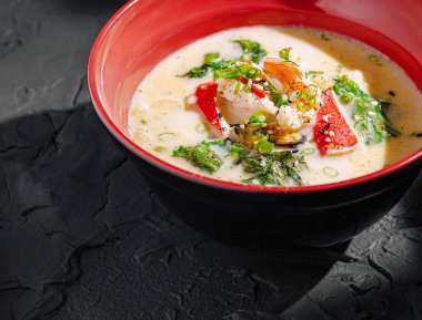 Karides, yeşillik ve dilimlenmiş biberle kaplı canlı bir kasede zengin bir deniz ürünleri çorbası servis edilir. Yemeklerde taze malzemeler ve canlı renkler sergileniyor. Lezzetli bir deneyim yaşanıyor..