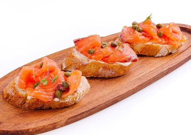 Füme somon, kapari ve otlarla kaplı taze pişmiş ekmek enfes bir aperatif sunar. Ahşap servis tabağında mükemmel bir şekilde yerleştirilmiş, bu tartinler hoşgörüye davet ediyor..