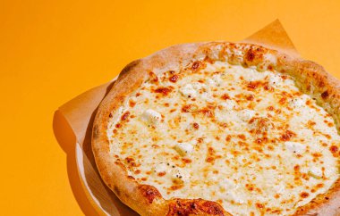 Taze bir pizza erimiş peynirle kaplanır ve yuvarlak tabakta altın bir kabuk sergilenir. Parlak turuncu arka plan bu klasik yemeğin iştah açıcı çekiciliğini arttırır..