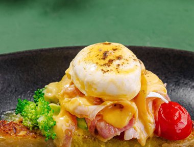 Ağız sulandıran bir kahvaltı tabağında leziz jambon ve erimiş peynirin üzerinde taze brokoli ve domateslerle çevrili haşlanmış bir yumurta bulunur. Siyah bir tabakta servis ediliyor, güne canlı bir başlangıç..