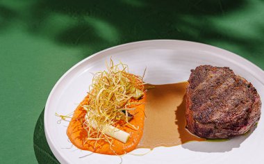 Renkli sebze püresi ve çıtır çıtır garnitürlerin yanında baharatlı bir et diliminin yer aldığı çok güzel sunulmuş bir biftek tabağı güzel bir yemek salonunda yemek pişirme sanatını gözler önüne seriyor..