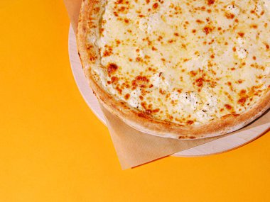 Altın kabuklu ve erimiş peynirli taze peynir pizzası ahşap servis tepsisinin üzerinde duruyor. Parlak sarı arka plan bu yemeğin iştah açıcı görünümünü arttırır..