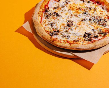 Yeni pişmiş bir pizza ahşap bir tahtanın üzerinde oturur, üzerinde erimiş peynir ve çeşitli lezzetli malzemelerle dolu altın bir kabuk sergilenir canlı bir portakal zeminine karşı.