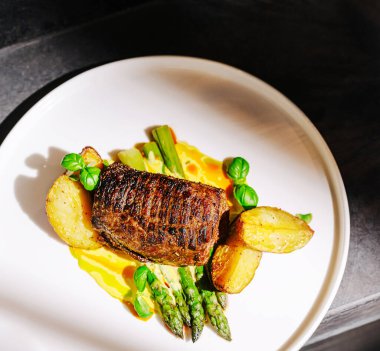 Kızarmış patates dilimleri ve taze kuşkonmazla birlikte bir tabak mükemmel ızgara biftek. Tabak canlı otlarla süslenmiş, mutfak sanatını gözler önüne seriyor..