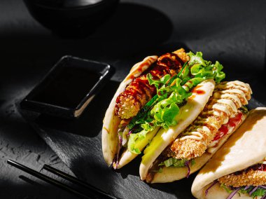 A set of three bao buns is filled with crispy chicken, fresh lettuce, and colorful vegetables on a dark serving board. A small dish of sauce and chopsticks are nearby. clipart