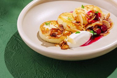 Üstü kızarmış hindistan cevizi, böğürtlen sosu ve kremayla kaplanmış üç altın sarısı krep. Tabak zarif bir şekilde açık renkli bir tabakta yeşil arka planda sunuluyor..