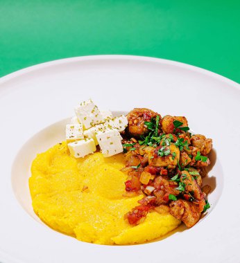 Lezzetli bir polenta tabağı sotelenmiş karides ve taze otlarla kaplanarak parlak yeşil arka planda sunulan renkli ve iştah açıcı bir yemek oluşturur..