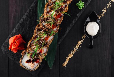 Tempura suşi rulosu, yanında wasabi, zencefil turşusu ve mayonez ile siyah bir tabakta servis ediliyor.
