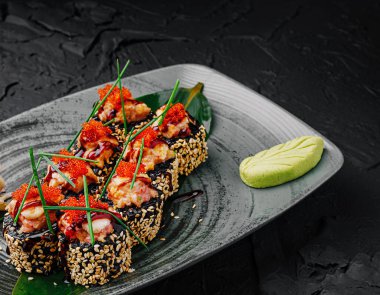Suşi dürümleri koyu bir tabakta güzelce düzenlenmiş, kırmızı balık yumurtası ve taze soğanla süslenmiş. Bir tutam wasabi rulonun yanında oturur, sunumu güçlendirir..