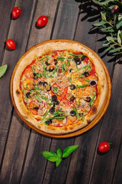 Taze sebzeler, siyah zeytinler ve otlarla süslenmiş renkli bir pizza kırsal bir ahşap masaya yerleştirilir. Çeri domatesleri ve yeşil fesleğen yaprakları tabağın etrafını sararak iştah açıcı bir ortam oluşturuyor..