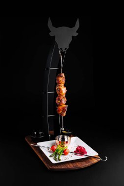 Yumuşak ızgara et şişleri kıvrımlı bir stantta zarif bir şekilde sergileniyor, canlı sebzeler ve soslarla birlikte, karanlık bir ortamda modern bir yemek deneyimi sergileniyor..