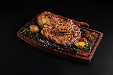 Büyük bir ızgara biftek ahşap bir tabakta zarif bir şekilde sunulur, yanında kızarmış patates ve otlarla süslenir. Bu yemek, et severler için ideal, lezzetli bir dokuya vurgu yapıyor..