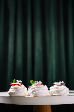 Çilek, yaban mersini ve nane aromalı üç zarif tatlı yuvarlak bir masaya oturur. Koyu yeşil bir perde bu zevkli gösteriye canlı bir zemin ekler.