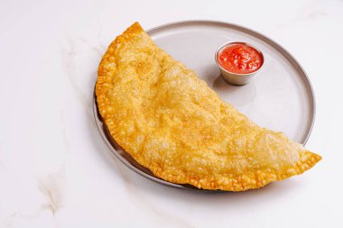 Bir altın kahverengi börek, küçük bir bardak baharatlı sosun yanında metal bir tabağın üzerinde duruyor. Yumuşak ışığın altında çıtır çıtır hamur işi parlıyor, dokusunu vurguluyor..