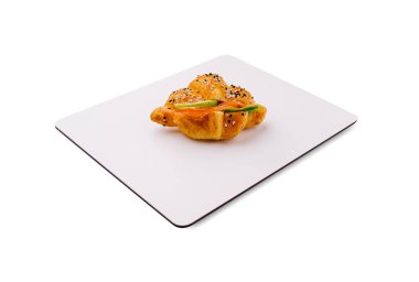 Sıcak bir kruvasan, susam tohumlarıyla kaplı ve taze malzemelerle dolu sade beyaz bir tabakta oturur. Dekor, pastanın tuhaf dokusunu ve iştah açıcı görünümünü ön plana çıkarıyor..