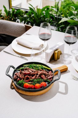 Güzel düzenlenmiş bir tabakta kırmızı şarap kadehleri ve şık bir ambiyansı olan zarif bir masaya kurulmuş canlı kiraz domatesleri ve taze yeşillikler bulunan yumuşak et dilimleri yer alıyor..